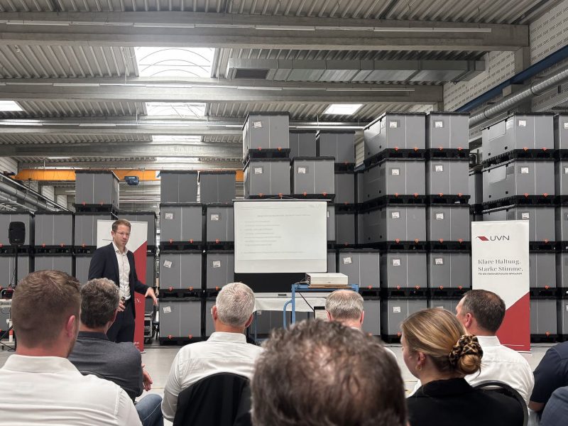 Foto vom Treffen des Rüstungsclusters Niedersachsen bei TKT Kunststoff-Technik GmbH. Es zeigt einen Redner, der in einer Fertigungshalle vor gestapelten Kisten einen Vortrag hält. Im Vordergrund sind einige Personen des Publikums von hinten zu sehen, die dem Vortrag zuhören.