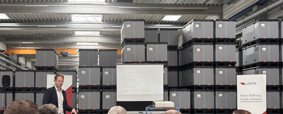 Foto vom Treffen des Rüstungsclusters Niedersachsen bei TKT Kunststoff-Technik GmbH. Es zeigt einen Redner, der in einer Fertigungshalle vor gestapelten Kisten einen Vortrag hält. Im Vordergrund sind einige Personen des Publikums von hinten zu sehen, die dem Vortrag zuhören.