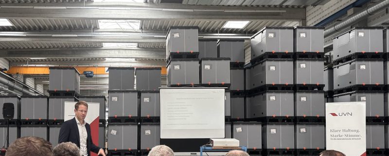 Foto vom Treffen des Rüstungsclusters Niedersachsen bei TKT Kunststoff-Technik GmbH. Es zeigt einen Redner, der in einer Fertigungshalle vor gestapelten Kisten einen Vortrag hält. Im Vordergrund sind einige Personen des Publikums von hinten zu sehen, die dem Vortrag zuhören.